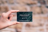 A hand is holding a small, dark green card with white text. The card features the words 'TERRES D'ÉMERAUDE' along with some symbols and a website URL at the bottom. The background is blurred, showing a mix of earthy tones.