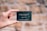 A hand is holding a small, dark green card with white text. The card features the words 'TERRES D'ÉMERAUDE' along with some symbols and a website URL at the bottom. The background is blurred, showing a mix of earthy tones.