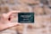 A hand is holding a small, dark green card with white text. The card features the words 'TERRES D'ÉMERAUDE' along with some symbols and a website URL at the bottom. The background is blurred, showing a mix of earthy tones.