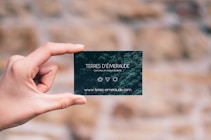 A hand is holding a small, dark green card with white text. The card features the words 'TERRES D'&Eacute;MERAUDE' along with some symbols and a website URL at the bottom. The background is blurred, showing a mix of earthy tones.