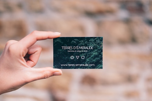 A hand is holding a small, dark green card with white text. The card features the words 'TERRES D'&Eacute;MERAUDE' along with some symbols and a website URL at the bottom. The background is blurred, showing a mix of earthy tones.