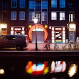 A nighttime street scene featuring a building with illuminated neon signs, including 'peep show' and colorful circular designs. In front of the building, there is a streetlamp casting light over a parked van and several bicycles. The scene is reflected in a canal running alongside the street.