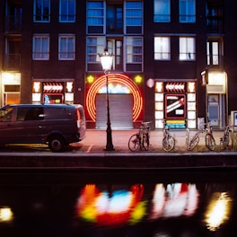 A nighttime street scene featuring a building with illuminated neon signs, including 'peep show' and colorful circular designs. In front of the building, there is a streetlamp casting light over a parked van and several bicycles. The scene is reflected in a canal running alongside the street.