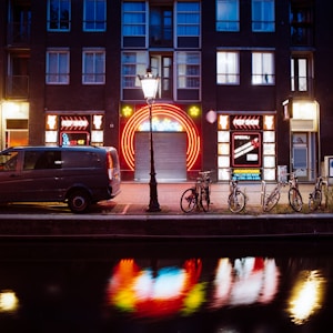 A nighttime street scene featuring a building with illuminated neon signs, including 'peep show' and colorful circular designs. In front of the building, there is a streetlamp casting light over a parked van and several bicycles. The scene is reflected in a canal running alongside the street.