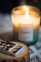 A lit candle labeled 'Forest' emits a warm glow in a glass container. In the foreground, a box of matches with blue tips and a floral design rests on a wooden coaster. The soft focus and warm lighting create a cozy atmosphere.