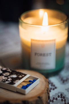 A lit candle labeled 'Forest' emits a warm glow in a glass container. In the foreground, a box of matches with blue tips and a floral design rests on a wooden coaster. The soft focus and warm lighting create a cozy atmosphere.