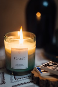 A lit candle labeled 'Forest' with scents of fennel, lemongrass, sandalwood, and mandarin is placed on a surface beside an open matchbox containing blue-tipped matches. The background is softly blurred, contributing to a warm and cozy ambiance.