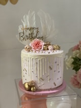 A beautifully decorated birthday cake with white and pink colors. It features gold accents and decorative macaron and rosette on the side. The cake has a 'Happy Birthday' topper with large white feathers behind it and is adorned with candy pearls and white icing drips.