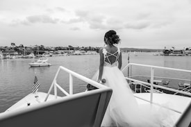A woman in a wedding dress stands on the deck of a boat looking out over a harbor. The background includes several moored boats and a distant shoreline with buildings. The weather appears overcast, and the scene is captured in black and white.