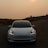 A sleek, modern white car is parked on a winding road under an overcast sky with the sun partially obscured by haze. The headlights are illuminated, and there are hills and a small dome-topped building visible in the background, creating a serene yet slightly mysterious atmosphere.