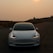 A sleek, modern white car is parked on a winding road under an overcast sky with the sun partially obscured by haze. The headlights are illuminated, and there are hills and a small dome-topped building visible in the background, creating a serene yet slightly mysterious atmosphere.