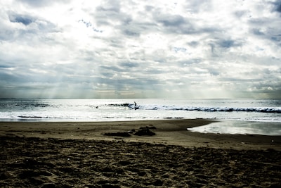 A serene beach scene with a sandy foreground and gentle waves lapping against the shore. In the distance, a lone surfer rides a wave under a dramatic sky filled with scattered clouds. Sunlight streams through gaps in the clouds, creating beautiful rays across the ocean.