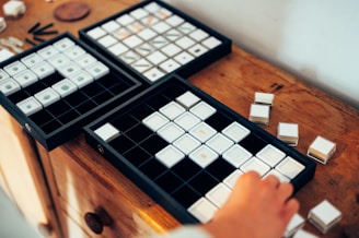 A wooden surface holds three open black trays, each containing an assortment of small, white, square tiles neatly arranged in grids. A hand is reaching towards one of the trays, suggesting interaction with the tiles. Several tiles are scattered on the table next to the trays.