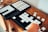 A wooden surface holds three open black trays, each containing an assortment of small, white, square tiles neatly arranged in grids. A hand is reaching towards one of the trays, suggesting interaction with the tiles. Several tiles are scattered on the table next to the trays.