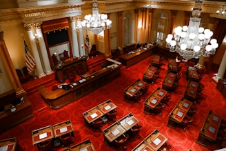 An ornate legislative chamber features rows of wooden desks and chairs placed over a richly patterned red carpet. The room is adorned with large chandeliers and elegant columns, with an American flag and another flag near the podium at the front. The high ceiling and intricate details add a sense of historical grandeur.