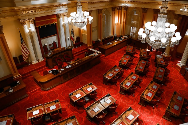 An ornate legislative chamber features rows of wooden desks and chairs placed over a richly patterned red carpet. The room is adorned with large chandeliers and elegant columns, with an American flag and another flag near the podium at the front. The high ceiling and intricate details add a sense of historical grandeur.