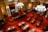 An ornate legislative chamber features rows of wooden desks and chairs placed over a richly patterned red carpet. The room is adorned with large chandeliers and elegant columns, with an American flag and another flag near the podium at the front. The high ceiling and intricate details add a sense of historical grandeur.