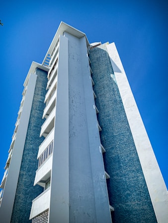 A tall, modern apartment building with a sleek design, featuring vertical lines and a combination of blue and white colors, set against a clear blue sky.