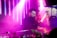 In a vibrant, dimly lit setting with pink and purple lighting, a DJ is seen focused on mixing music while a woman beside him captures the moment with her smartphone. The atmosphere is lively and energetic.