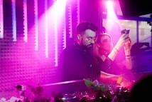 In a vibrant, dimly lit setting with pink and purple lighting, a DJ is seen focused on mixing music while a woman beside him captures the moment with her smartphone. The atmosphere is lively and energetic.