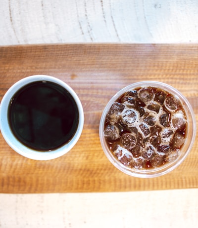 Two beverages are placed on a wooden surface, with one being a cup of hot black coffee and the other a glass of iced coffee filled with ice cubes.
