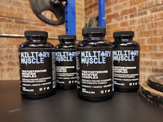 Four bottles labeled 'Military Muscle Testosterone Booster Complex' are arranged on a dark surface. The background consists of a brick wall and gym equipment, including weight plates.