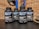 Four bottles labeled 'Military Muscle Testosterone Booster Complex' are arranged on a dark surface. The background consists of a brick wall and gym equipment, including weight plates.