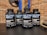 Four bottles labeled 'Military Muscle Testosterone Booster Complex' are arranged on a dark surface. The background consists of a brick wall and gym equipment, including weight plates.