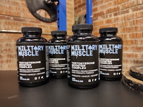 Four bottles labeled 'Military Muscle Testosterone Booster Complex' are arranged on a dark surface. The background consists of a brick wall and gym equipment, including weight plates.