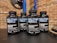 Four bottles labeled 'Military Muscle Testosterone Booster Complex' are arranged on a dark surface. The background consists of a brick wall and gym equipment, including weight plates.