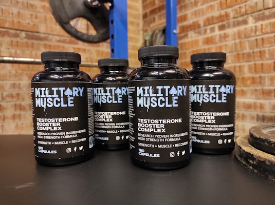 Four bottles labeled 'Military Muscle Testosterone Booster Complex' are arranged on a dark surface. The background consists of a brick wall and gym equipment, including weight plates.
