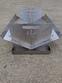 A metallic, foldable structure with serrated edges and a central cut-out design, placed on a concrete surface. The structure appears to have handles and symmetrical sides.