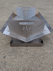 A metallic, foldable structure with serrated edges and a central cut-out design, placed on a concrete surface. The structure appears to have handles and symmetrical sides.
