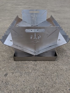 A metallic, foldable structure with serrated edges and a central cut-out design, placed on a concrete surface. The structure appears to have handles and symmetrical sides.