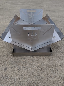 A metallic, foldable structure with serrated edges and a central cut-out design, placed on a concrete surface. The structure appears to have handles and symmetrical sides.