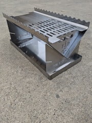 A metal, portable fireplace unit with a grated top and serrated edges sits on a concrete surface. The unit appears sleek and well-constructed, designed for outdoor use.
