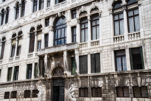 A grand, historic facade of a building featuring multiple arched windows with intricate grillwork, detailed stone carvings, and statues. The structure is predominantly made of light-colored stone, and the windows have a repetitive pattern with both clear and dark finishes.