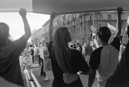 A black and white image depicts a street protest with a crowd of people marching and holding flags. In the foreground, three individuals are holding a large banner above their heads. The scene is set in an urban environment with buildings lining the street.
