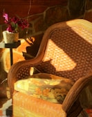 A sunny porch with a wicker chair wrapped in a chunky knit throw blanket, surrounded by green plants and fresh flowers.