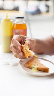 A hand holding a piece of food with visible layers of ham and cheese, accompanied by a drizzle of a creamy yellow sauce. On the table, a plate holds a folded crepe with sauce, along with a bottle of orange juice and a yellow squeeze bottle likely containing mustard or a similar condiment.