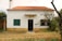 A small, rustic house with a red-tiled roof and white walls. The front features green shuttered windows, a central door, and a chimney. The garden is overgrown with some trees and plants, giving it a slightly neglected appearance.