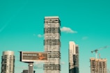 A cityscape featuring modern high-rise buildings under construction, with a prominent billboard advertisement in the foreground. A construction crane is visible, indicating urban development. The sky is a clear turquoise, adding a striking backdrop to the scene.
