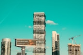A cityscape featuring modern high-rise buildings under construction, with a prominent billboard advertisement in the foreground. A construction crane is visible, indicating urban development. The sky is a clear turquoise, adding a striking backdrop to the scene.