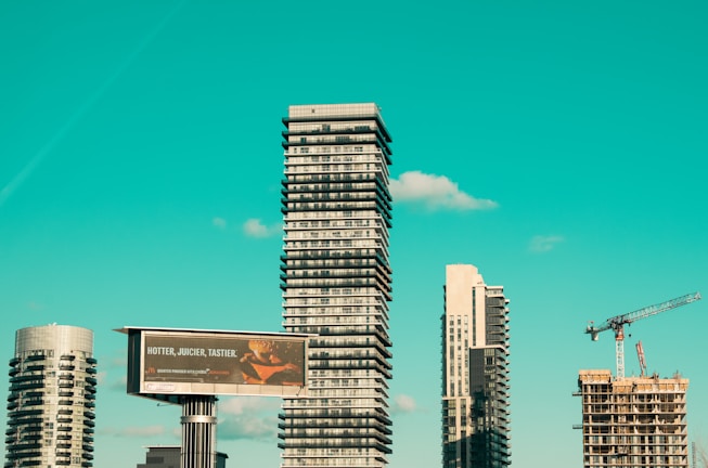 A cityscape featuring modern high-rise buildings under construction, with a prominent billboard advertisement in the foreground. A construction crane is visible, indicating urban development. The sky is a clear turquoise, adding a striking backdrop to the scene.