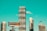A cityscape featuring modern high-rise buildings under construction, with a prominent billboard advertisement in the foreground. A construction crane is visible, indicating urban development. The sky is a clear turquoise, adding a striking backdrop to the scene.