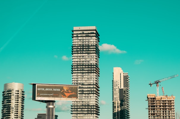 A cityscape featuring modern high-rise buildings under construction, with a prominent billboard advertisement in the foreground. A construction crane is visible, indicating urban development. The sky is a clear turquoise, adding a striking backdrop to the scene.