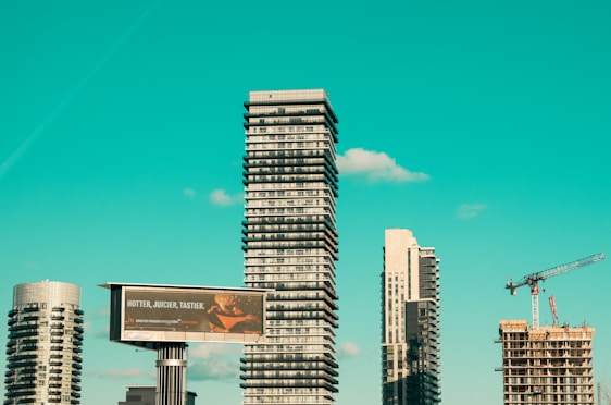 A cityscape featuring modern high-rise buildings under construction, with a prominent billboard advertisement in the foreground. A construction crane is visible, indicating urban development. The sky is a clear turquoise, adding a striking backdrop to the scene.