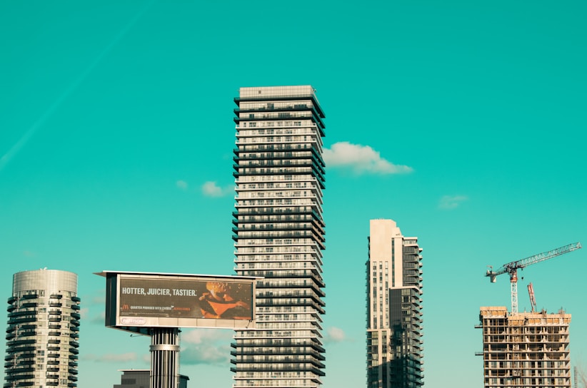 A cityscape featuring modern high-rise buildings under construction, with a prominent billboard advertisement in the foreground. A construction crane is visible, indicating urban development. The sky is a clear turquoise, adding a striking backdrop to the scene.