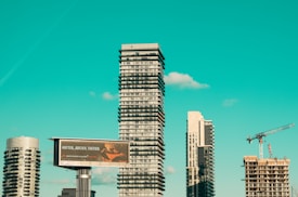 A cityscape featuring modern high-rise buildings under construction, with a prominent billboard advertisement in the foreground. A construction crane is visible, indicating urban development. The sky is a clear turquoise, adding a striking backdrop to the scene.
