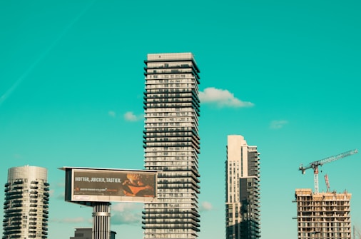 A cityscape featuring modern high-rise buildings under construction, with a prominent billboard advertisement in the foreground. A construction crane is visible, indicating urban development. The sky is a clear turquoise, adding a striking backdrop to the scene.